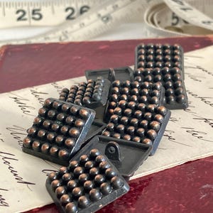 以下が含まれることがあります： 小さな銅色の球体のテクスチャ表面を持つ、正方形の濃い灰色のボタンのコレクション。ボタンは、手書きのスクリプトが書かれたヴィンテージ紙の上に配置され、背景にはメジャーテープがあります。