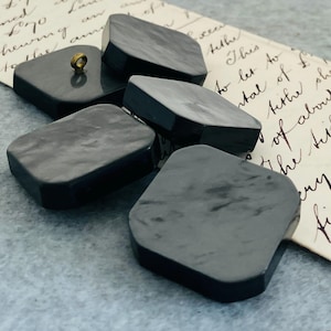 May include: Five dark grey, square buttons with rounded corners. One button has a gold-coloured shank. The buttons are arranged on a light blue surface with a piece of vintage paper with handwritten text.