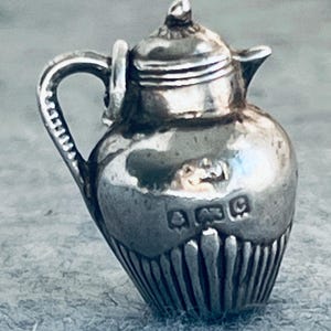 Könnte beinhalten: Ein silberner Miniatur-Teekannen-Anhänger mit bauchigem Körper, vertikalen Rillen am Boden, einem geschwungenen Henkel, einem kleinen Ausguss und einem Deckel. Auf der Vorderseite sind drei deutliche Punzierungen in das Metall eingeprägt.