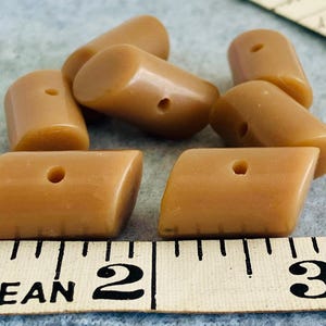 Puede incluir: Un conjunto de cuentas en forma de barril de color marrón claro, con un agujero perforado en cada una. Las cuentas están dispuestas sobre una superficie azul claro junto a una cinta métrica. Las cuentas miden aproximadamente 2,5 cm de largo.