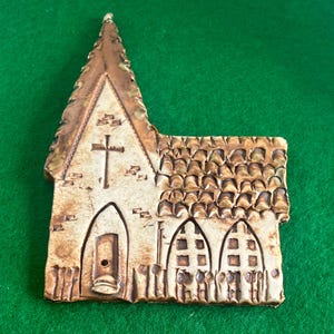 May include: A flat, ceramic wall plaque shaped like a church building. The piece features a tall steeple, a central cross, a single arched doorway, and two arched windows. The surface is finished in a cream colour with brown, glazed accents.