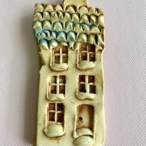 May include: A small ceramic house ornament with a blue and white tiled roof, three windows, and a door. The house is painted in a light beige color and has a rustic, handmade look.