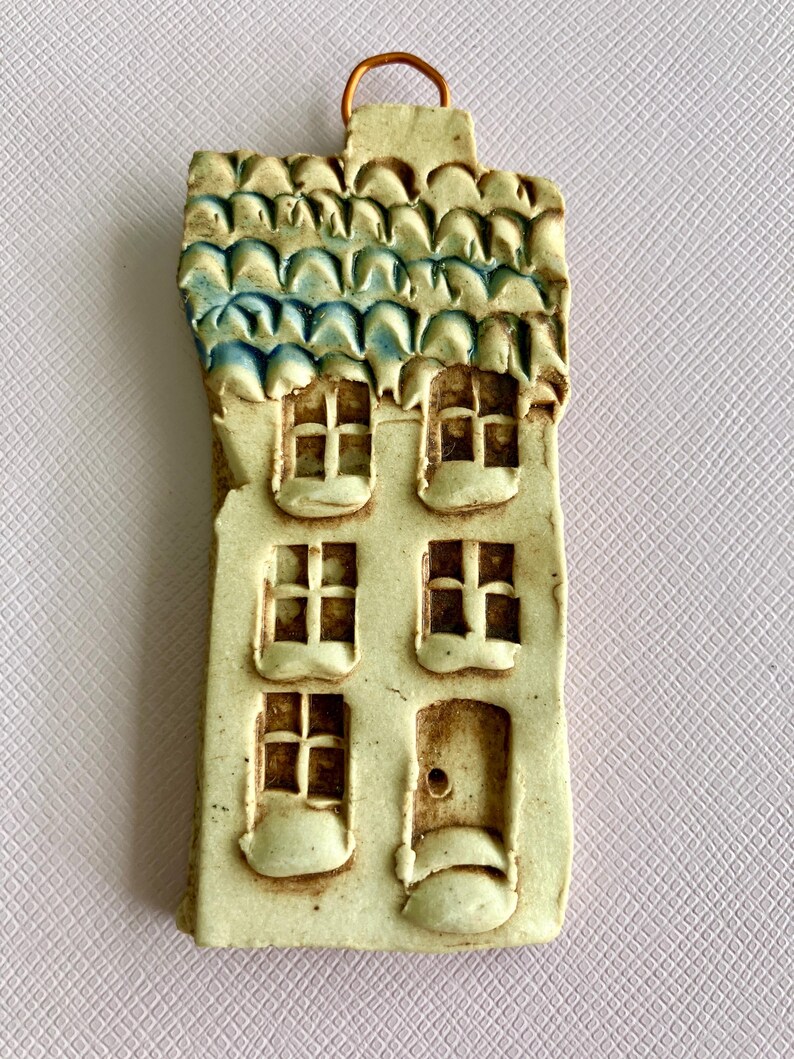 May include: A small ceramic house ornament with a blue and white tiled roof, three windows, and a door. The house is painted in a light beige color and has a rustic, handmade look.