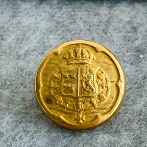 May include: A circular gold button with an embossed heraldic design. The button features a crown above a shield with detailed imagery. The button has a vintage appearance, with signs of wear. The button's diameter is approximately 2.5 cm.