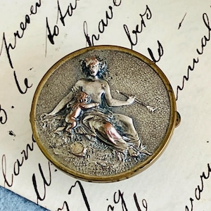 May include: A circular, antique silver-coloured compact featuring a detailed relief of a seated woman and child. The compact has a gold-coloured rim, set against a backdrop of handwritten script.