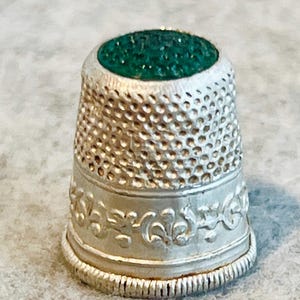 May include: A silver-toned metal thimble featuring a textured, dimpled surface across the upper portion and a decorative embossed band with scrolling patterns near the base. The top of the thimble is inlaid with a circular, dark green stone.