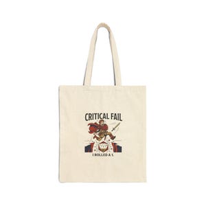 May include: A cream-coloured tote bag with a design featuring a cartoon figure in a red cape, a sword, and the text "CRITICAL FAIL" and "I ROLLED A 1." The bag has two handles and is made of a sturdy fabric.