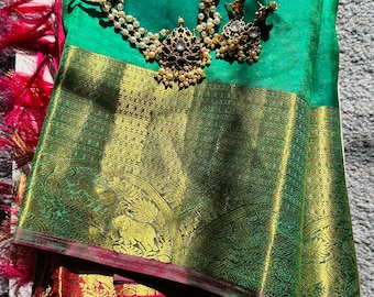 May include: A green and gold silk saree with a red border. The saree is folded and draped on a white surface. A pearl and gold necklace and earrings are on top of the saree.