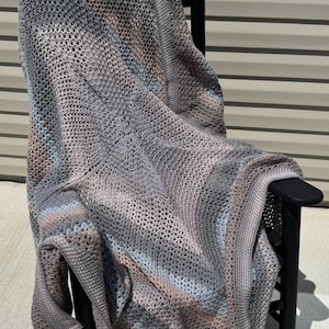 May include: A hand-crocheted blanket featuring a variegated yarn pattern in shades of gray, soft blue, and muted pink. The textile displays a consistent open-stitch texture throughout, with a finished border edge draped over a black wooden chair.