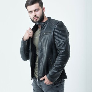 Peut inclure: Un homme portant une veste en cuir noire avec une fermeture &eacute;clair et un t-shirt vert en dessous. Il porte un jean bleu avec des d&eacute;tails us&eacute;s.