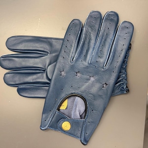 Puede incluir: Un par de guantes de conducción de cuero azul marino. Los guantes tienen recortes en forma de estrella y pequeñas perforaciones para ventilación. Un botón dorado con la letra "A" está en la muñeca.