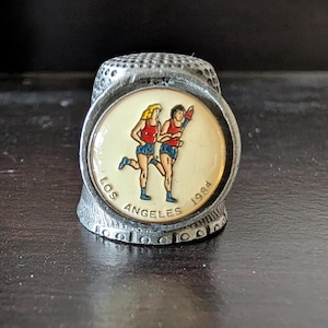 May include: A silver thimble with a white and gold circular design featuring two runners holding a torch. The design also includes the text "Los Angeles 1984".