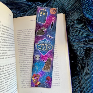 Puede incluir: Un marcapáginas colorido de Doctor Who con el TARDIS, un Dalek y otros elementos icónicos sobre un fondo estrellado. El marcapáginas está colocado en un libro abierto.