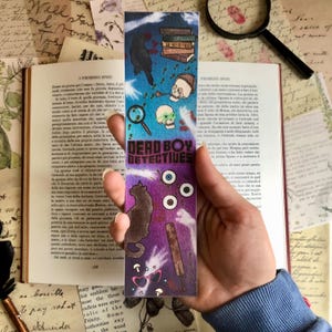May include: A purple and blue bookmark with a black cat, a crow, a magnifying glass, a skull, and a baseball bat. The bookmark has the text "Dead Boy Detectives" in white letters.