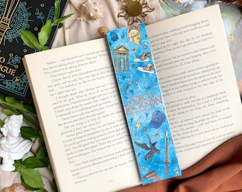May include: A blue and white bookmark featuring a character from the Percy Jackson series. The bookmark has a blue background with a white border and features a drawing of a character with a blue baseball cap, a sword, and a winged creature. The text "Percy Jackson and the Olympians" is printed on the bookmark.