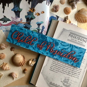 Op de afbeelding: Een blauwe en turquoise bladwijzer met de tekst "Child of Poseidon" in rood-oranje. De bladwijzer heeft een aquarelachtig ontwerp met een golfpatroon. De tekst "Poseidone Dio del Mare" is zichtbaar op de achtergrond.