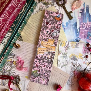 May include: A pink bookmark with a floral pattern and the text "C'era una volta...". The bookmark features illustrations of a pumpkin, a frog, and other whimsical elements. Antique keys and vintage books surround the bookmark.