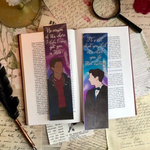 May include: Two colorful bookmarks with quotes from the show "The Umbrella Academy." The left bookmark features a blue and purple background with a silhouette of a man wearing a jacket and the quote "No version of this where I didn't come get you, is there?" The right bookmark features a blue and green background with a silhouette of a man wearing a suit and the quote "It's not what you did, it's what you do that matters."