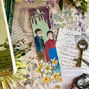 Può includere: Un segnalibro rettangolare in carta con un'illustrazione colorata di due figure in piedi tra narcisi gialli sotto fiori di glicine viola. Il segnalibro mostra una persona con una giacca blu e un'altra con un cappotto rosso, entrambe con gli occhiali.