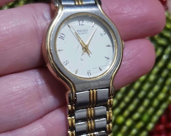 May include: A two-tone silver and gold watch with a white face and black hands. The watch face has black Roman numeral markings and the words "SEIKO QUARTZ" printed on it.
