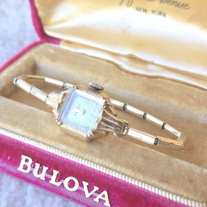May include: A gold-tone Bulova wristwatch with a rectangular face and a matching link bracelet, presented in a pink and cream-colored velvet-lined display box with the text Bulova Fifth Avenue New York printed on the interior lid.