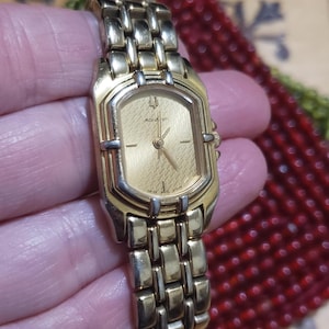 May include: Gold-toned Accutron watch with a rectangular face and a matching link bracelet. The watch face is gold with black hands and markers. The watch is held in a hand, with a blurred background.