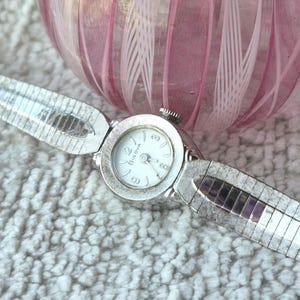 May include: A silver-tone Bulova wristwatch featuring a round white dial with silver numerals and hands. The watch has a textured silver metal expansion band with a polished finish. The watch case is circular with a small crown at the top position.
