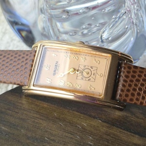 May include: A rectangular gold-tone Gruen Curvex wristwatch with a light brown textured leather strap. The watch face features gold-colored Arabic numerals, gold hands, and a small square sub-dial for seconds positioned above the six o'clock marker.