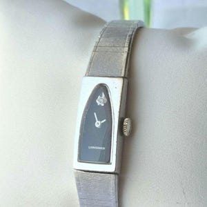May include: A silver-toned Longines wristwatch featuring a unique teardrop-shaped case with a black dial, a single diamond accent at the twelve o'clock position, silver hands, and a flexible, brushed metal link bracelet with a side winding crown.