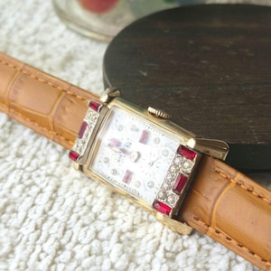 May include: A rectangular gold-tone wristwatch with a white dial featuring the text "WHITELY". The watch case is accented with clear stones and rectangular red stones at the top and bottom. It has a tan leather strap with a crocodile-embossed texture.