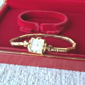 May include: A gold-toned wristwatch with a square face and a white dial featuring black numerals. The watch has a slender, articulated metal link bracelet and a small winding crown on the side. It is presented in a red velvet-lined display case.