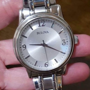 May include: A silver Bulova watch with a white face and silver hands. The watch has a silver metal band.