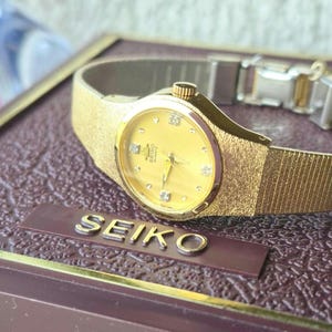 May include: A gold-tone Seiko Quartz wristwatch featuring a round yellow dial with small crystal hour markers, gold hands, and a textured gold-tone metal link bracelet. The watch is displayed on a dark brown box with the brand name SEIKO in gold.