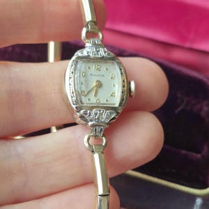 May include: A vintage Bulova wristwatch featuring a silver-toned rectangular case with intricate filigree detailing at the top and bottom lugs. The off-white dial displays gold-toned numerals at the 12, 2, 4, 6, 8, and 10 positions with matching hands.