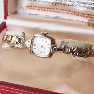 May include: A gold-tone Wittnauer wristwatch with a square face, white dial, and gold-tone markers, featuring a textured link bracelet. The watch is presented in a red and cream-colored box with the text Wittnauer A Longines-Wittnauer Product.