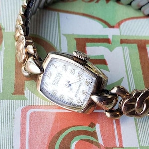 May include: A gold-toned Gruen Curvex wristwatch with a white face and black Roman numeral hour markers. The watch has a gold-toned metal band with a unique link design.
