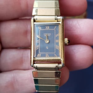 May include: A gold-toned Citizen Quartz watch with a rectangular face and black Roman numeral hour markers. The watch has a gold-toned metal band.