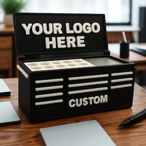 以下が含まれることがあります： 蓋に「YOUR LOGO HERE」と白く書かれた黒いツールボックス型のオーガナイザー。オーガナイザーには、9つの小さな円形のコンパートメントと、「CUSTOM」と白く書かれたラベルの付いた引き出しが2つあります。