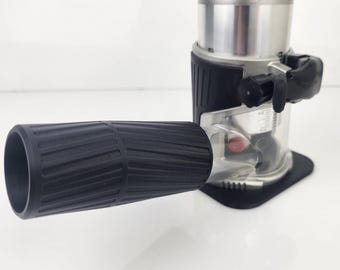 May include: A close-up of a power tool, a router, with a black handle and a silver and black body. The router has a clear base and a black grip. The tool is on a white surface.