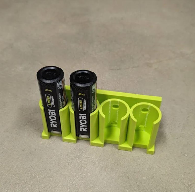 May include: A lime green 3D-printed battery holder with two black and yellow Ryobi USB Lithium batteries inserted. The holder has three slots for batteries and is designed to keep them organized.
