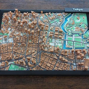 May include: A 3D relief map of Tokyo, Japan, in a black frame. The map features detailed representations of buildings, streets, and green spaces, with the city name displayed at the top. The map is in shades of brown, green, and blue.