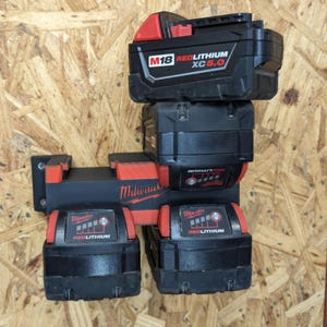 May include: Black and red Milwaukee M18 Redlithium XC 5.0 battery pack and charger mount. The batteries are stacked on top of each other and mounted on a wooden surface. The charger mount is black and red.