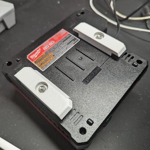 May include: A black Milwaukee power tool battery charger with a white plastic latch on the side. The charger has a warning label that reads "WARNING!" in red letters. The label also includes the text "ADVERTENCIA!" in Spanish.