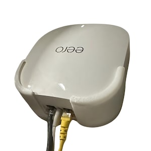 Op de afbeelding: Een witte Eero Wi-Fi-router met twee ethernet-kabels aangesloten. De router is aan een muur gemonteerd.