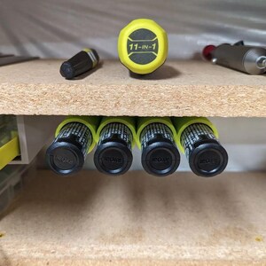 May include: A yellow and black 11-in-1 screwdriver with a yellow handle and black text. Four additional screwdriver bits are stored below. The tools are on a wooden shelf.
