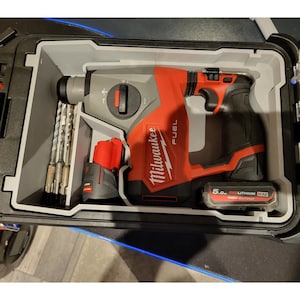 Könnte beinhalten: Ein rotes und graues Milwaukee FUEL Hammerbohrerset in einem grauen Koffer. Das Set enthält den Bohrer, einen Akku und Bohrer. Der Bohrer hat einen schwarzen Griff und das Milwaukee-Logo. Der Koffer ist geöffnet und zeigt die Werkzeuge.
