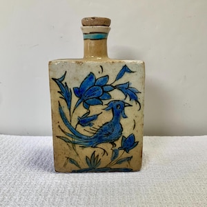 May include: A rectangular ceramic bottle with a beige speckled glaze and a cork stopper. The front features a hand-painted blue bird surrounded by blue floral motifs. The neck of the bottle has a horizontal blue band near the top opening.