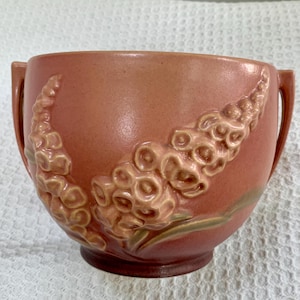 May include: A peach-colored ceramic bowl with two handles and a floral relief design. The bowl has a textured surface and a dark brown base. The floral design is in a lighter peach color with green accents.