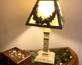 May include: An antique table lamp with a square shade, adorned with floral detailing, and a decorative base. The lamp sits on a table next to a wooden box with floral inlay. The lamp's shade is approximately 12 inches wide.