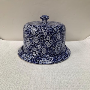May include: A blue and white ceramic cheese dome with a floral pattern. The dome has a rounded top with a small knob and sits on a matching plate. The floral design covers the entire piece.